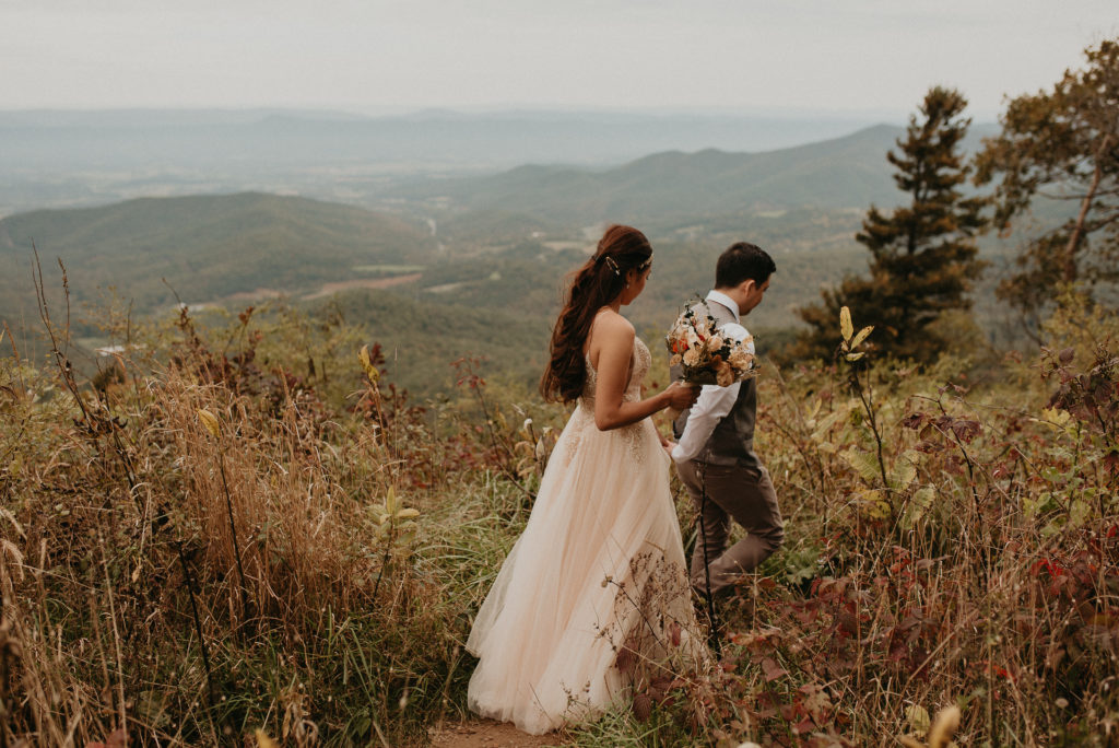 Groom leading his bride down a trail on the side of the mountain in Shenandoah National Park. Adventurous hiking elopement in Virginia. Wooden bridal bouquet.