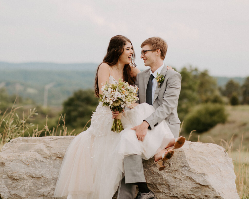 bride and groom laughing on a rock overlooking the blue ridge mountains in the summer